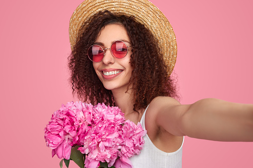 Charming curly haired female in straw hat and sunglasses taking self shot with bunch of peony flowers while looking at camera on pink background