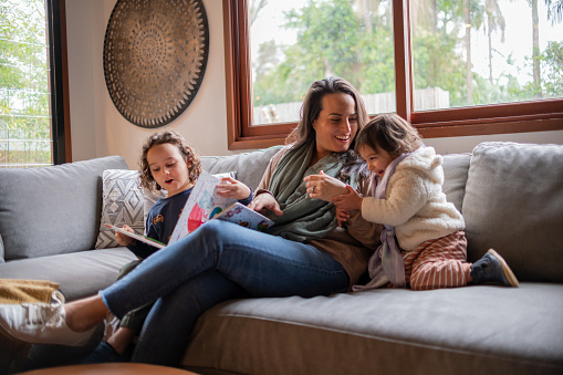 A mother laughs and plays with her two young children. They are snuggled up on the couch together and enjoying quality family time.