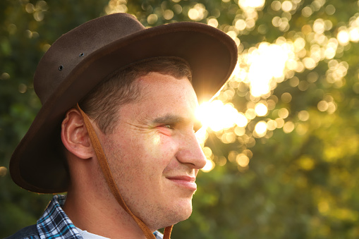 Young man farmer in cowboy hat at agricultural field on green blurred background. Closeup portrait of millennial man with hat, standing on nature background, outdoors. Rancher. Sunset.