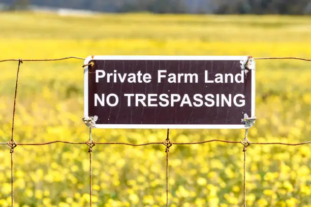 Private Farm Land No Trespassing sign posted on a barbed wire fence. Close up. Blurred yellow mustard field.