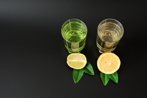 Two transparent glasses with citrus juices on a black background, next to half a ripe lemon and lime with leaves. Top view, flat lay.