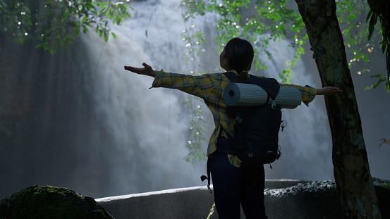 Rear View Asian young women with backpacks raise their arms into the air while open arms enjoying the view of a waterfall, 24-year-old woman, Trekking, Adventure, Freedom, Happy, holiday, independence, inspiration, joy, Freshness. Video Still