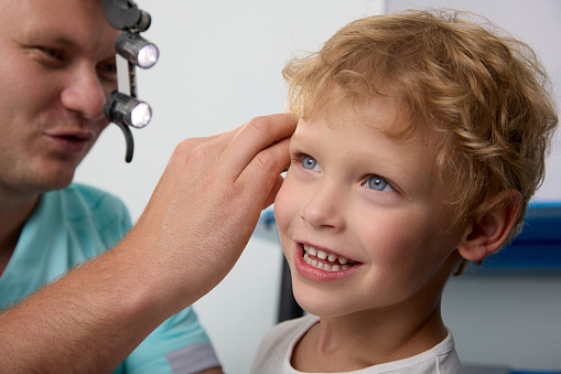 Otolaryngologist checks the hearing of a blond boy in a children's hospital, uses a headlamp. Reception and consultation of an experienced ENT doctor of a little boy smiling during the examination