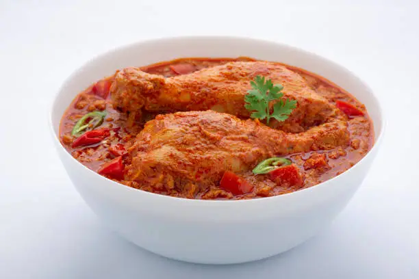 Chicken curry or masala , spicy reddish chicken leg piece dish garnished with coriander leaf and  fresh green chilli which is arranged in a white ceramic bowl with white background,isolated.