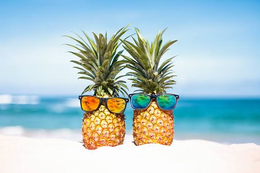 Couple of attractive pineapples in stylish mirrored sunglasses on the sand against turquoise sea. Tropical summer vacation concept