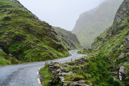 Narrow winding asphalt road wet with rain climbing among lush green rocky hills of Ring of Kerry in southwestern Ireland with misty sky opening in background with copy space