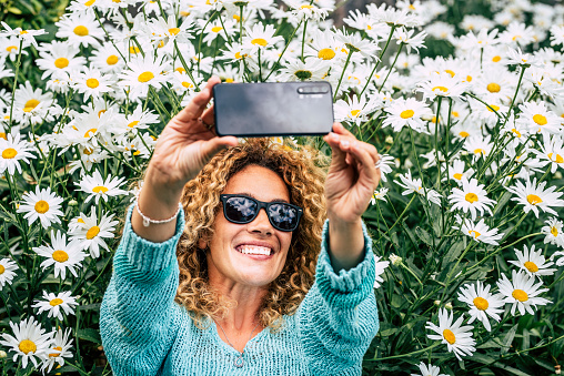 Springtime. Welcome spring. Happy woman above view taking selfie picture laying on the grass with a lot of daisy blossom flowers around. Sharing portrait on social media concept modern online life