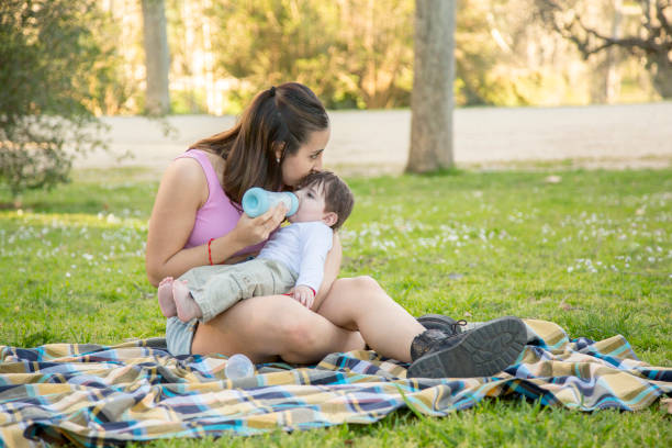 jeune nouvelle mère assise sur la pelouse embrassant son bébé tout en nourrissant son bébé au biberon - single mother one parent child kissing photos et images de collection