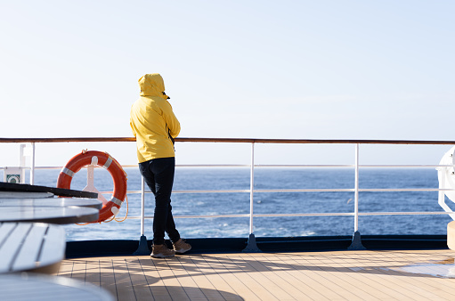 A cruise passenger in a yellow parka stands at the railing at the stern of a cruise ship and looks back at the blue open sea.  Next to the person hangs an orange life preserver.
