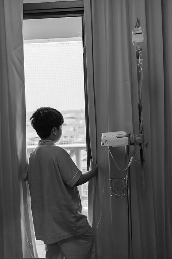 Asian boy admitted in hospital while saline intravenous (IV) on hand. Unhappy boy depressed and hopeless with absent minded looking outside.