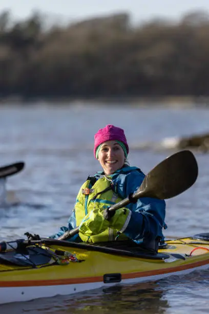 A front-view close up shot of a mid adult woman kayaking in the sea, she is paddling with an oar and she is wearing a warm hat and life jacket.