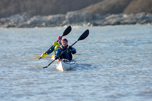 A front-view shot of two women kayaking in the sea, they are paddling with their oars and they are wearing life jackets, enjoying travelling on the calm waters.