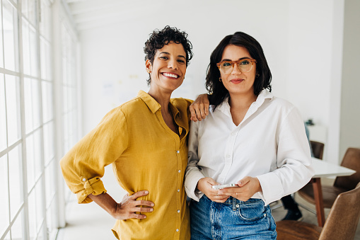 Portrait of two business women standing together in an office. Female colleagues looking at the camera. Women working in a multicultural workplace.