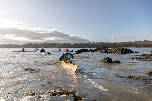 A wide-view shot of a small group of mature and mid adult friends kayaking in the sea, they are paddling with oars and they are wearing life jackets.