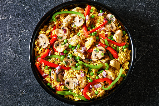chicken mushroom paella with sweet bell pepper, green peas and spices in black bowl on concrete table, horizontal view from above, flat lay, close-up