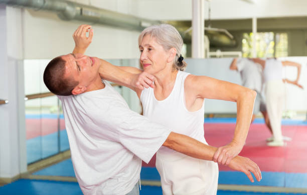 Aged woman practicing elbow blow with wristlock to opponent in gym Determined aged woman learning self defence techniques in sparring with young man, practicing elbow blow with wristlock to opponent in gym self defense stock pictures, royalty-free photos & images