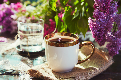 Coffee break with cup of coffee among lilac flowers. Aesthetic cottage core lifestyle on sunny day.