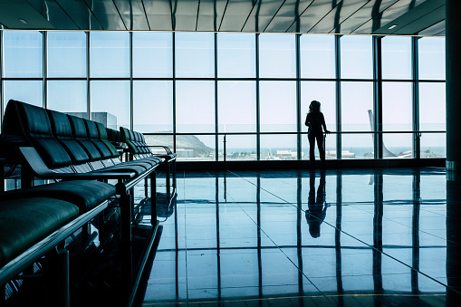 Travel concept. Back view of woman passenger in silhouette waiting her flight at the gate. Blue modern light mood. Tourist wait to fly. Business or vacation trip. Enjoying airplane to transport