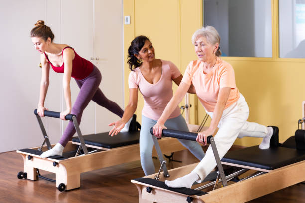 elderly woman practicing pilates on reformer with hispanic female trainer - senior adult physical therapy gymnastics cheerful imagens e fotografias de stock