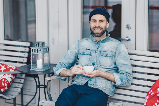 People, rest and positiveness concept. Smiling delighted bearded male model with happy expression rests in cozy cafe, holds mug with tea, being in high spirit, thinks about something pleasant