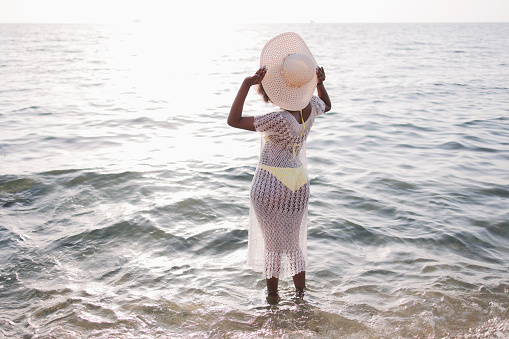 African-american woman in a swimsuit and beach dress stands tall in the sea or ocean and holds on to her hat. Summer mood, tropical, sea view, relaxing,rest in hot countries, ocean, waves, sunset