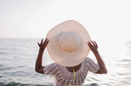 Woman in a hat, standing with her hands behind her hat on the beach, enjoys the summer holidays on a sunny summer day. Summertime, holding a white hat, holiday, leaisure, lifestyle, activity