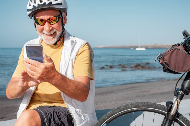 istock Cheerful active senior man with bicycle on a sunny day sits at the beach using mobile phone. Authentic retirement life and healthy lifestyle concept. Horizon over the sea 1482778752