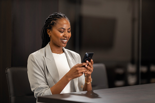 A beautiful black businesswoman is using a smartphone while waiting for a late meeting in the conference room.