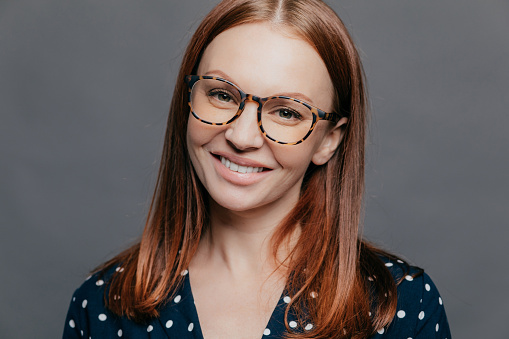 Cheerful close up shot of pretty woman with brown hair, toothy smile, wears spectacles, dressed in formal blouse, poses over grey background, being pleased to meet with colleagues on meeting