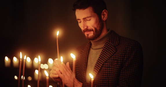 Aesthetic Shot of Young Christian Man Lighting a Candle in Church, Praying and Expressing Devotion to the Lord. Parishioner Being Enlightened by the Teachings of Jesus Christ and Following his Path