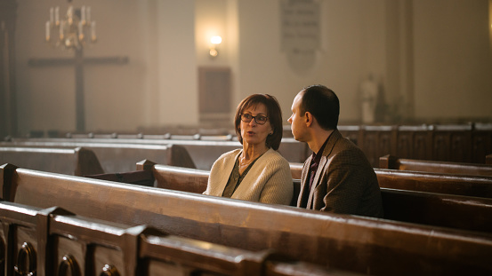 Portrait of Young Man and Old Woman Talking and Discussing While Sitting in a Church. Faithful Parishioners Seeking Comfort In Faith And Religious Beliefs, Consulting, Sharing Experiences and Wisdom