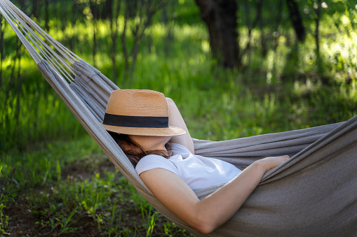 Woman resting in a hammock in a summer garden covering her face with a straw hat. Summer vacation concept