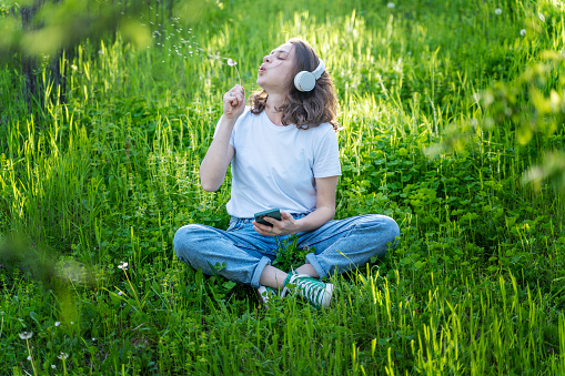 Young happy woman listening to music with headphones on a spring meadow blowing on dandelions