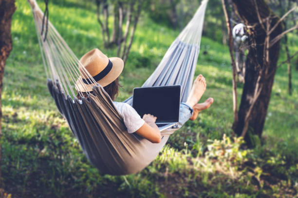istock Young woman in a hat using a laptop while lying in a hammock in a summer garden. Summer lifestyle concept, working online 1482789010