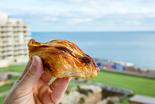 Local Maltese food called pastizz or pastizzi. Person hand holding Diamond-shaped small pie with Malta Sliema city on background with blue sky and sea.