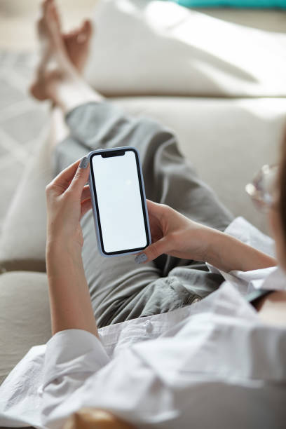 istock Mock up white screen blank mobile phone in woman hands holding lying on the sofa Back view 1482797041