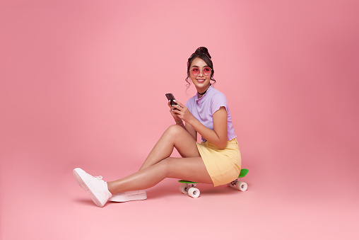 Full length happy young asian woman girl using smartphone and sitting on skateboard isolated on pastel pink background. People lifestyle concept. Sit on skateboard