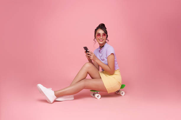 full length happy young asian woman girl using smartphone and sitting on skateboard isolated on pastel pink background. people lifestyle concept. sit on skateboard - women young adult isolated length imagens e fotografias de stock