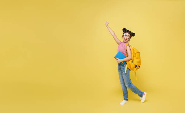 黄色い背景に全身の笑顔の幸せな陽気なアジアの学生女の子がバックパックの本を持つ。学校構想における教育 - child student adolescence cute ストックフォトと画像
