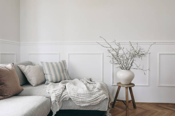 istock Stylish living room with vase and blooming cherry plum tree branches. Springtime home decor. Elegant scandinavian interior with comfy sofa, linen cushions and blanket. White wall background, nobody. 1482799760