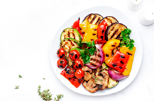 Grilled vegetables: paprika, zucchini, eggplant, mushrooms, tomatoes and onion on plate, white table background, top view
