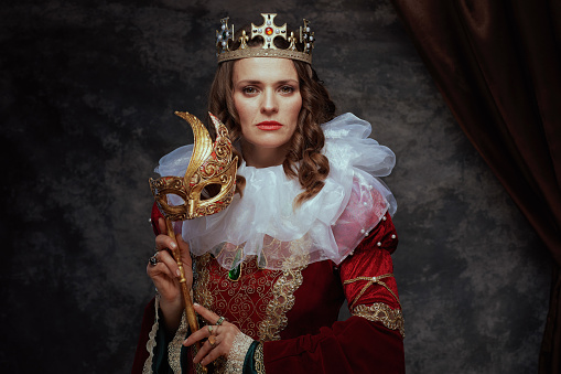 medieval queen in red dress with venetian mask, white collar and crown on dark gray background.