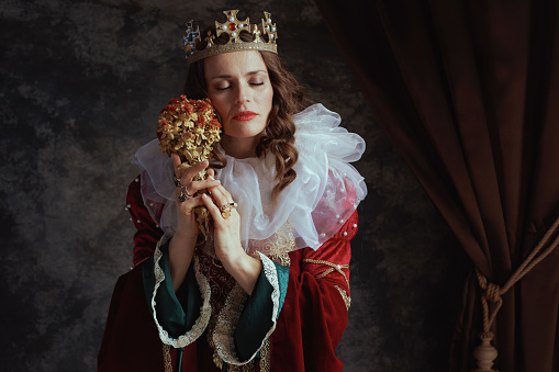 medieval queen in red dress with dried flower, white collar and crown on dark gray background.