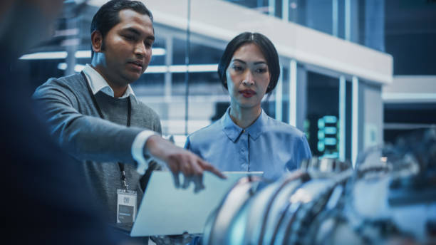 Two Multiethnic Employees Work in Modern Innovative Research Facility with a Futuristic Jet Engine. Engineer with a Laptop Explaining How the Prototype Motor Works to an Industrial Specialist. dos empleados multiétnicos trabajan en modernas instalaciones de investigación innovadoras con un motor a reacción futurista. ingeniero con una computadora portátil explicando cómo funciona el prototipo de motor a un especialista industrial.