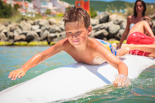 Cheerful teenage boy lying on a swimming board. Happy european family having fun floating on a swim board in the sea on a hot summer day