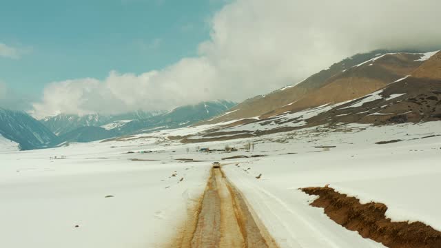 Rear view of a white car driving on a dirt road next to a roadside covered with snow