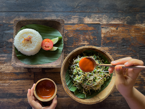 Traditional Indonesian vegan bowlof long green beans, spinach and bean sprouts served with rice and sambal sauce.  Zero waste leaf, wood plate, bowl, cup and table.  Ubud, Bali, Indonesia.