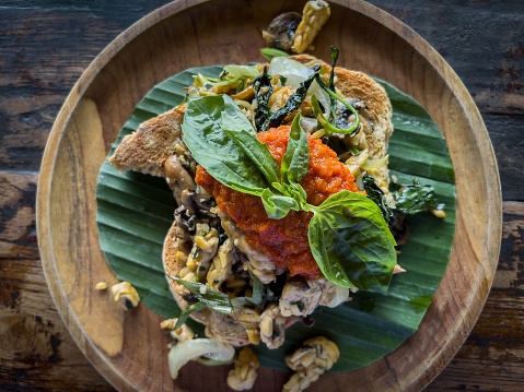 Indonesian-Western fusion breakfast: toast with tempeh, spicy tomato sauce, grilled vegetables, garnished with basil leaves.  Zero waste leaf, wood plate and table.  Ubud, Bali, Indonesia.