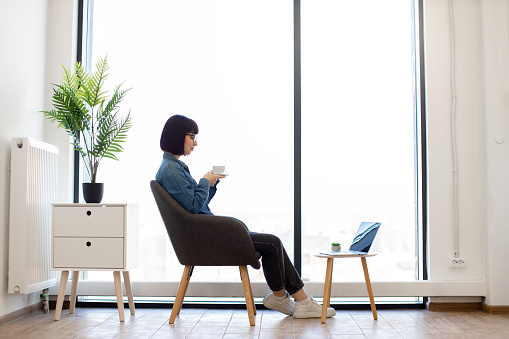 Dark haired woman in casual wear and glasses sitting in office armchair near panoramic window and enjoying hot aroma coffee. Small coffee table with portable laptop standing in front of female worker.