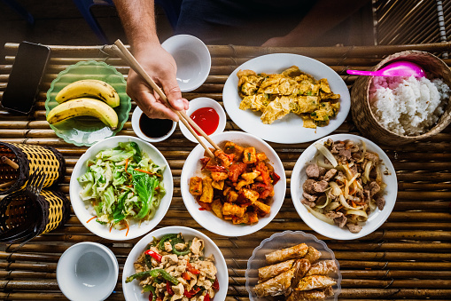 overhead view on male hand picking meat with chop sticks at table with various vietnamese dishes